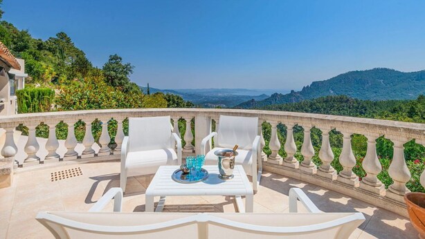 A terrace with white chairs and a table, a decorative railing, and a view of hills and greenery.
