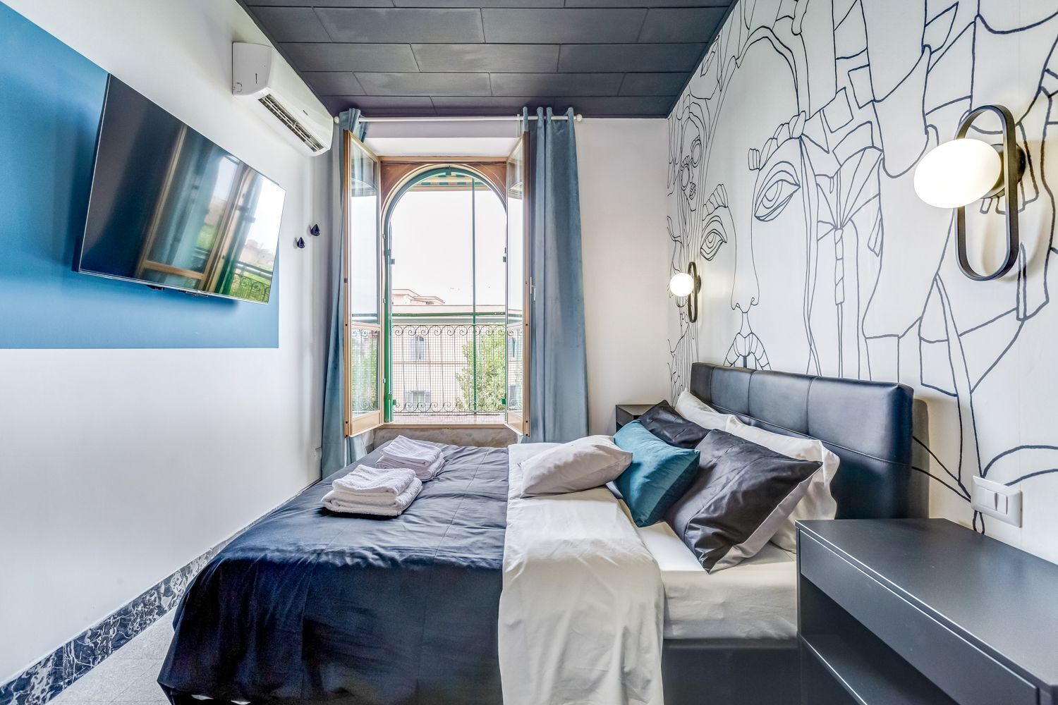 A modern bedroom with a bed, a flat-screen TV mounted on the wall, a window with curtains, and a decorative wall mural.
