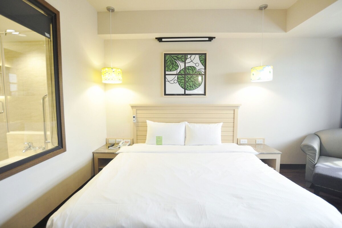 A hotel room with a large bed, bedside lamps, a nightstand with a phone, a chair, and a framed artwork on the wall.