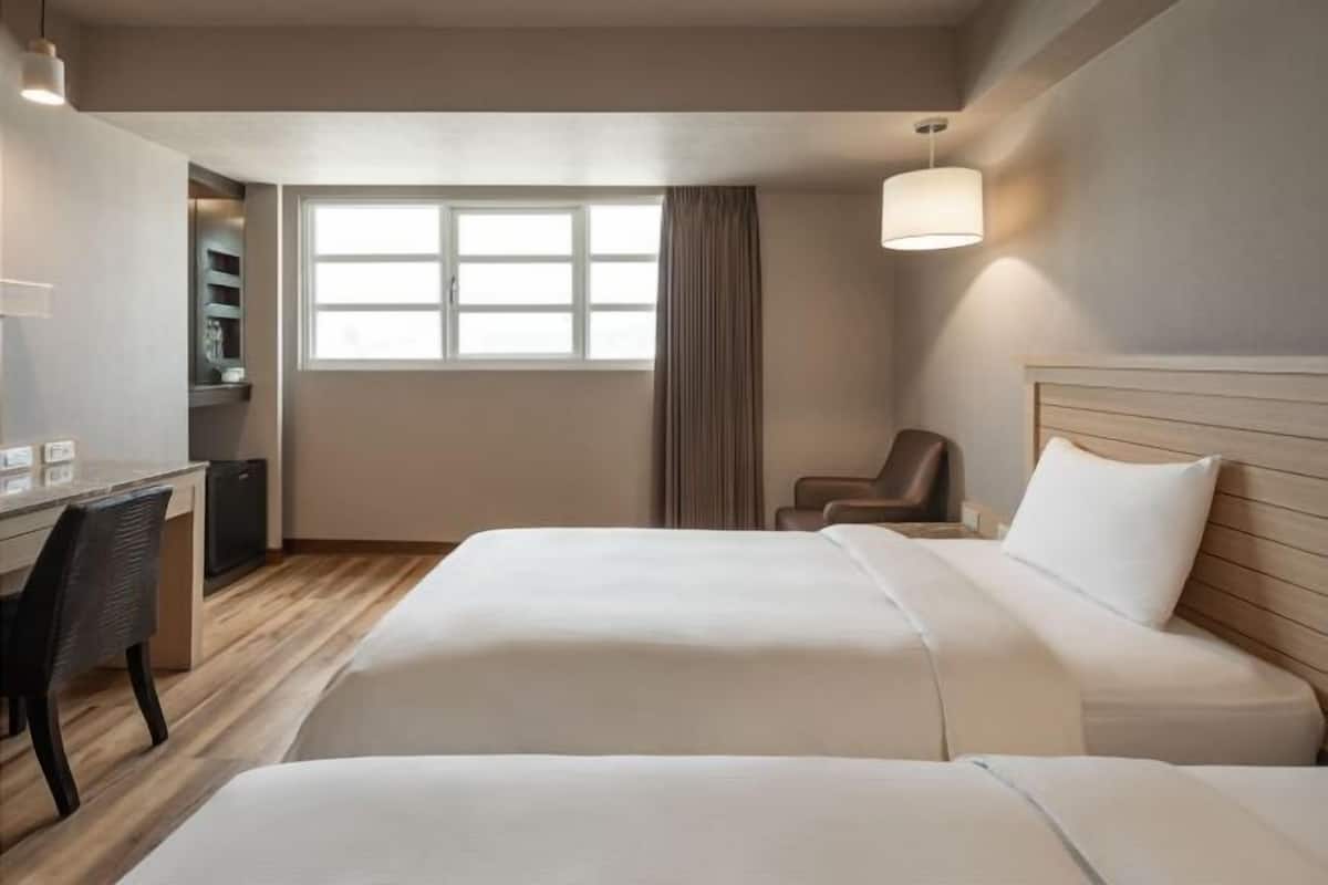 A hotel room with two beds, a desk, a chair, a lamp, and a window with curtains.