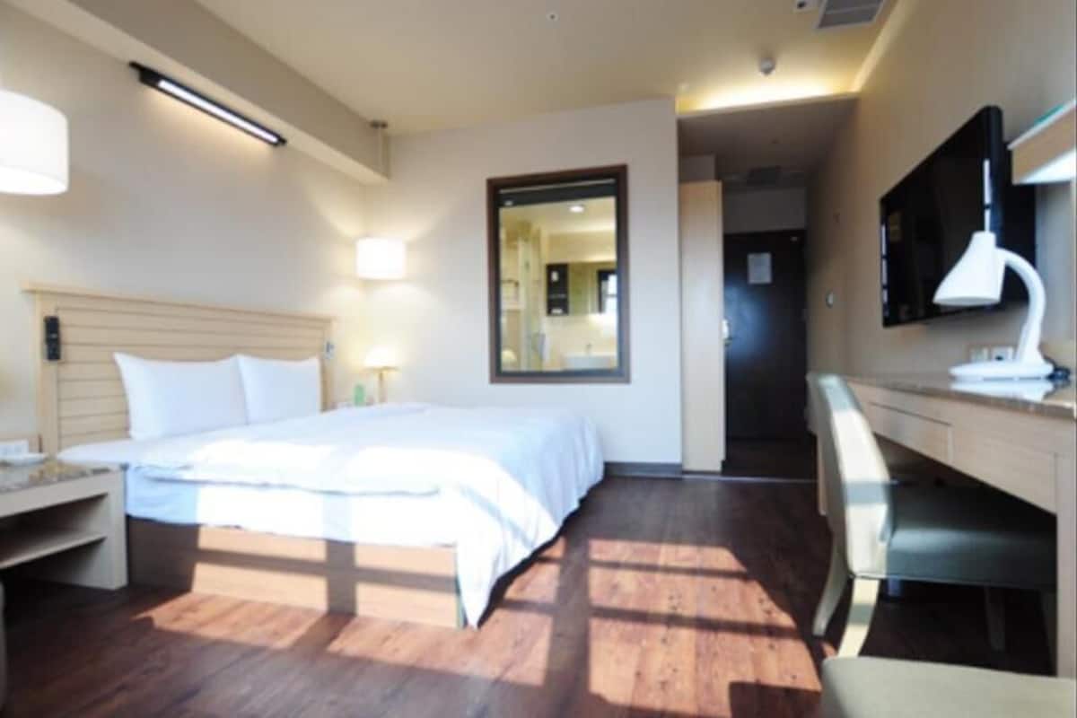 A hotel room with a bed, desk, chair, and a mirror on the wall.