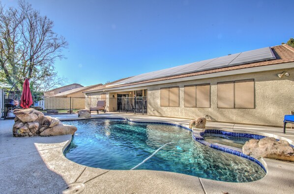 A swimming pool with a curved edge, a covered patio area, and a building with a tiled roof in the background.