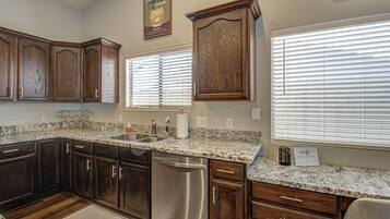 A kitchen with dark wooden cabinets, a stainless steel dishwasher, granite countertops, and a window with blinds.