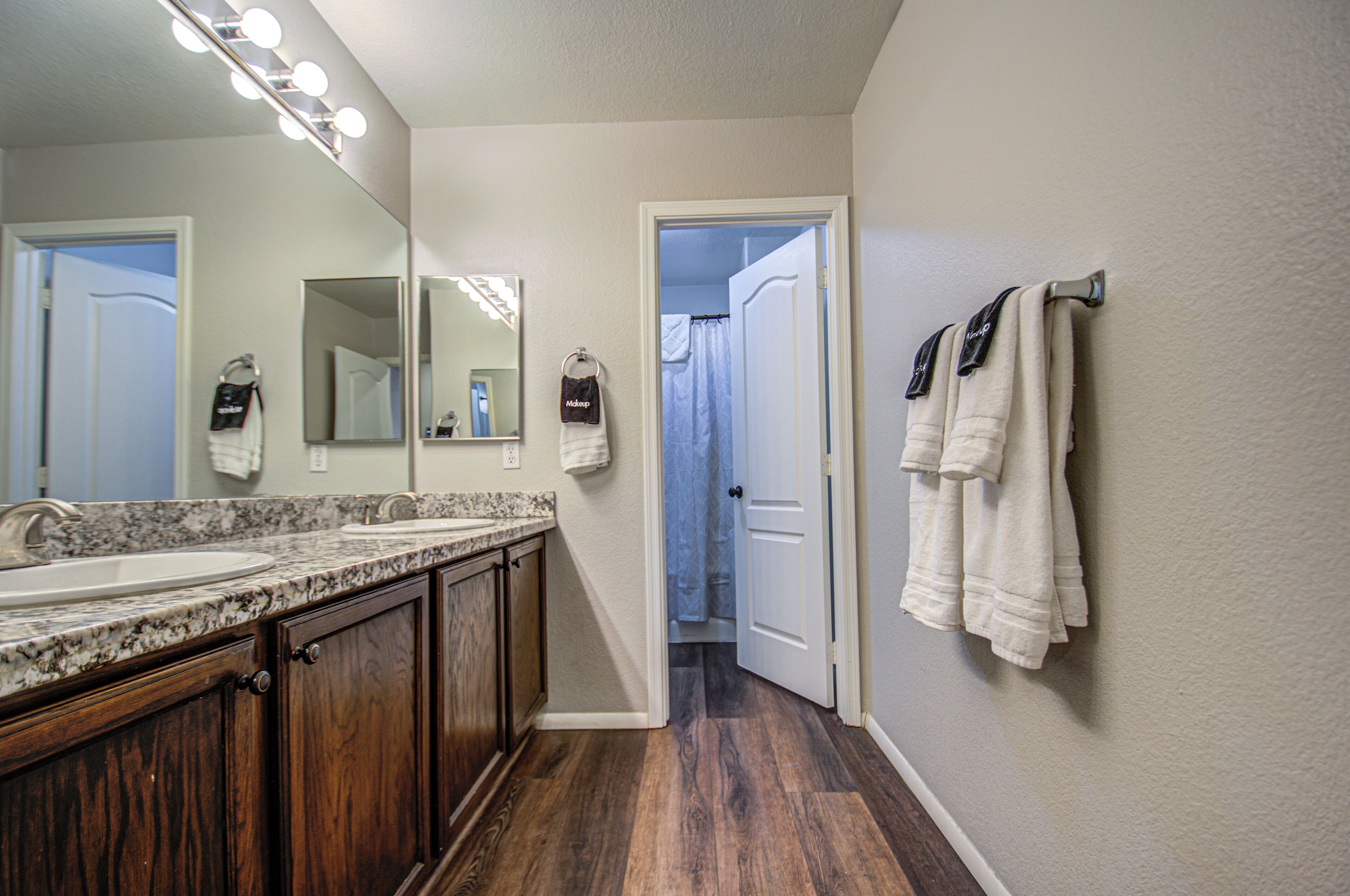 A bathroom with a double sink, wooden cabinets, a large mirror with lights, and a towel rack with towels.
