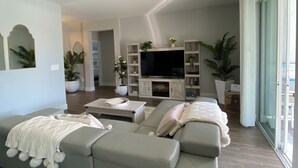A modern living room with a grey sofa, a flat-screen TV, and a large window.