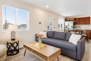 Ein modernes Wohnzimmer mit einem grauen Sofa, einem Holztisch und einer Blumenvase. Es gibt eine Küchenzeile mit Holdekor-Schränken und einem Kühlschrank.