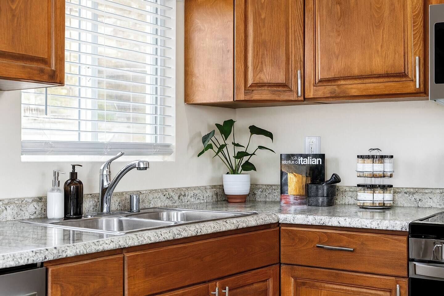 A kitchen with wooden cabinets, a granite countertop, a stainless steel sink, a potted plant, and a cookbook on the counter.