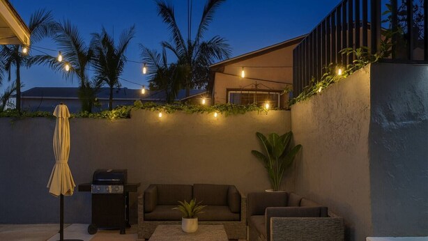 A modern outdoor seating area with a sofa, coffee table, and umbrella, surrounded by greenery and string lights.