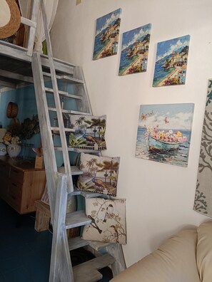 A room with a wooden ladder leading to a loft, a wooden desk with decorative items, and multiple wall-mounted paintings depicting coastal scenes.