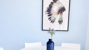 Una mesa redonda blanca con cuatro sillas blancas, un jarrón azul con flores moradas y un cuadro en la pared con la imagen de un pájaro.