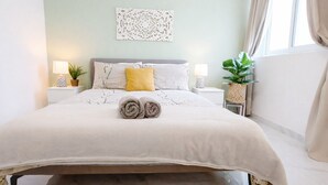 Una cama bien hecha con toallas enrolladas, un adorno de pared y dos mesitas de noche con lámparas y plantas.