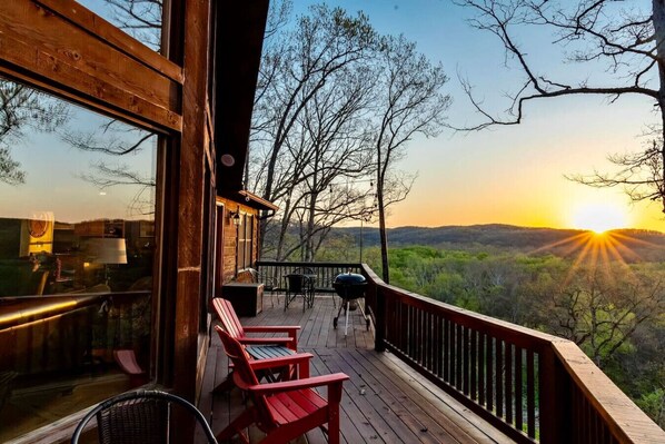 Eine Holzterrasse mit roten Adirondack-Stühlen und einem Grill, mit Blick auf eine bewaldete Landschaft bei Sonnenuntergang.
