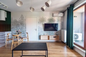 A modern living room with a black coffee table, wooden chairs, a TV on a wooden stand, and a wall with a nature-themed mural. - Waterlane Island Deluxe 4 by GrandApartm (Gdansk)
