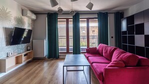 Ein Wohnzimmer mit rotem Sofa, einem an der Wand montierten Fernseher, einem Holztisch und Blick auf eine Stadtkulisse durch große Fenster.