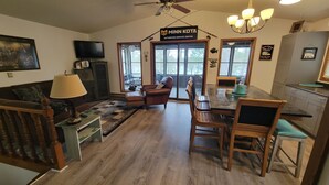 A living room with a sofa, a coffee table, a dining table with chairs, a TV mounted on the wall, and a sign indicating a service center. -  Newly listed: Ray's Retreat  (Webster)