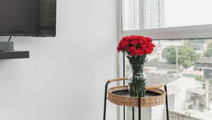 A room with a TV on a wall, a wicker chair with a vase of red flowers, and a view of buildings through a window.