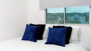 A bed with white bedding and two dark blue pillows, a window with a view of buildings, and a light-colored wall.