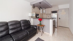 A modern living room with a black leather sofa, a small wooden side table with a vase of red flowers, and a kitchen area with a microwave, coffee maker, and refrigerator.