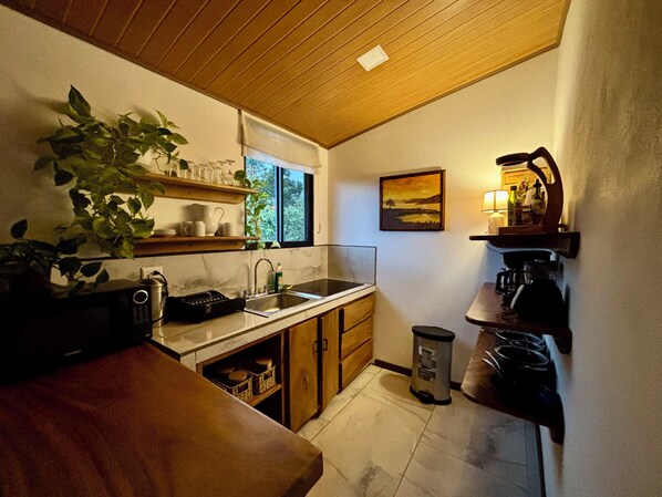 A compact kitchen with wooden cabinets, a sink, a window with blinds, a trash bin, and a shelf with kitchen items.