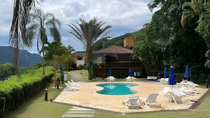 A pool area with lounge chairs and umbrellas, surrounded by greenery and a building in the background. - Linda casa em Boiçucanga (Sao Sebastiao)