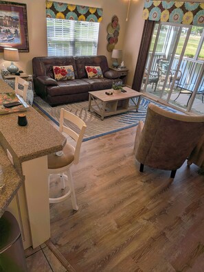 Una sala de estar con un sofá de cuero marrón, una mesa de centro de madera, una silla blanca y una zona de cocina con encimera de granito.