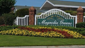 A sign for Magnolia Greens Golf Plantation with a white picket fence and a landscaped flower bed in front.