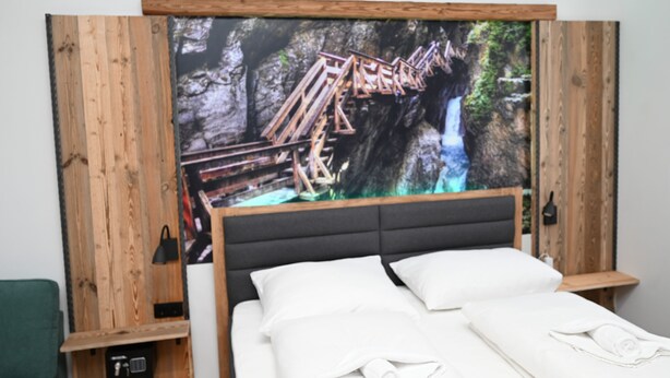 Een bed met een houten hoofdbord, een televisie erboven met daarop een uitzicht op een waterval, en een klein plankje met een zwarte lamp.