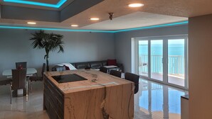 Una cocina moderna con una isla de mármol, zona de comedor y vistas al mar a través de amplios ventanales correderas.