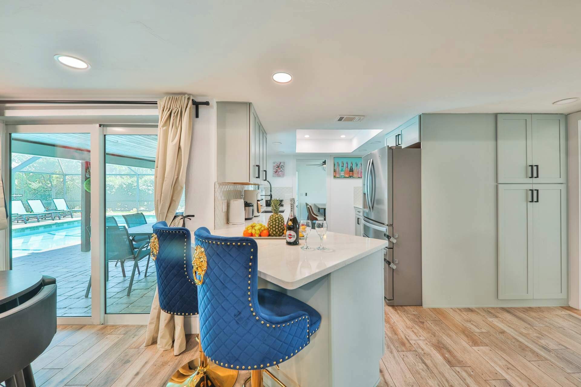 A modern kitchen with a large island, a dining area with blue chairs, and a view of a pool through a glass door.