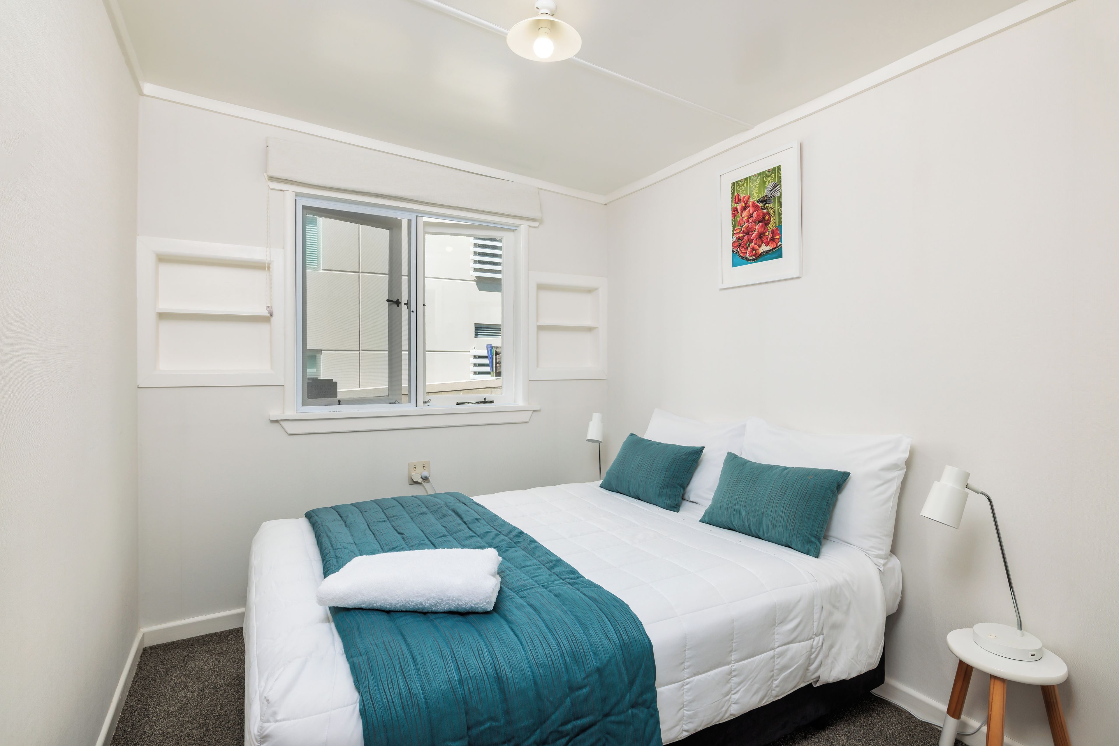 Une chambre à coucher avec un lit, une fenêtre, une table de chevet, une lampe et un tableau accroché au mur.