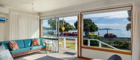 A spacious living room with a sofa, ottoman, and a small table. A large sliding glass door leads to a balcony with a view of a lake and parked cars.