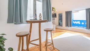 Una stanza moderna con un tavolo in legno e due sgabelli, una grande finestra con tende e vista su un muro blu e un balcone.