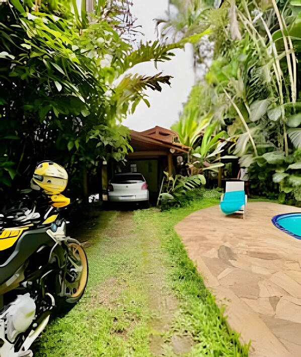 Una motocicleta estacionada en un jardín exuberante con un coche al fondo, una piscina y un pabellón.