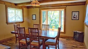 Une salle à manger avec une table et des chaises en bois, de grandes fenêtres donnant sur la forêt et un lustre.