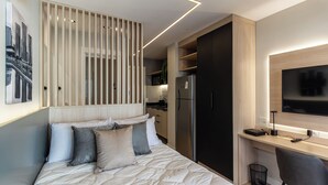 A modern bedroom with a bed, a desk, a chair, a TV, and a kitchen area. - 360 Oscar Itaim (São Paulo)