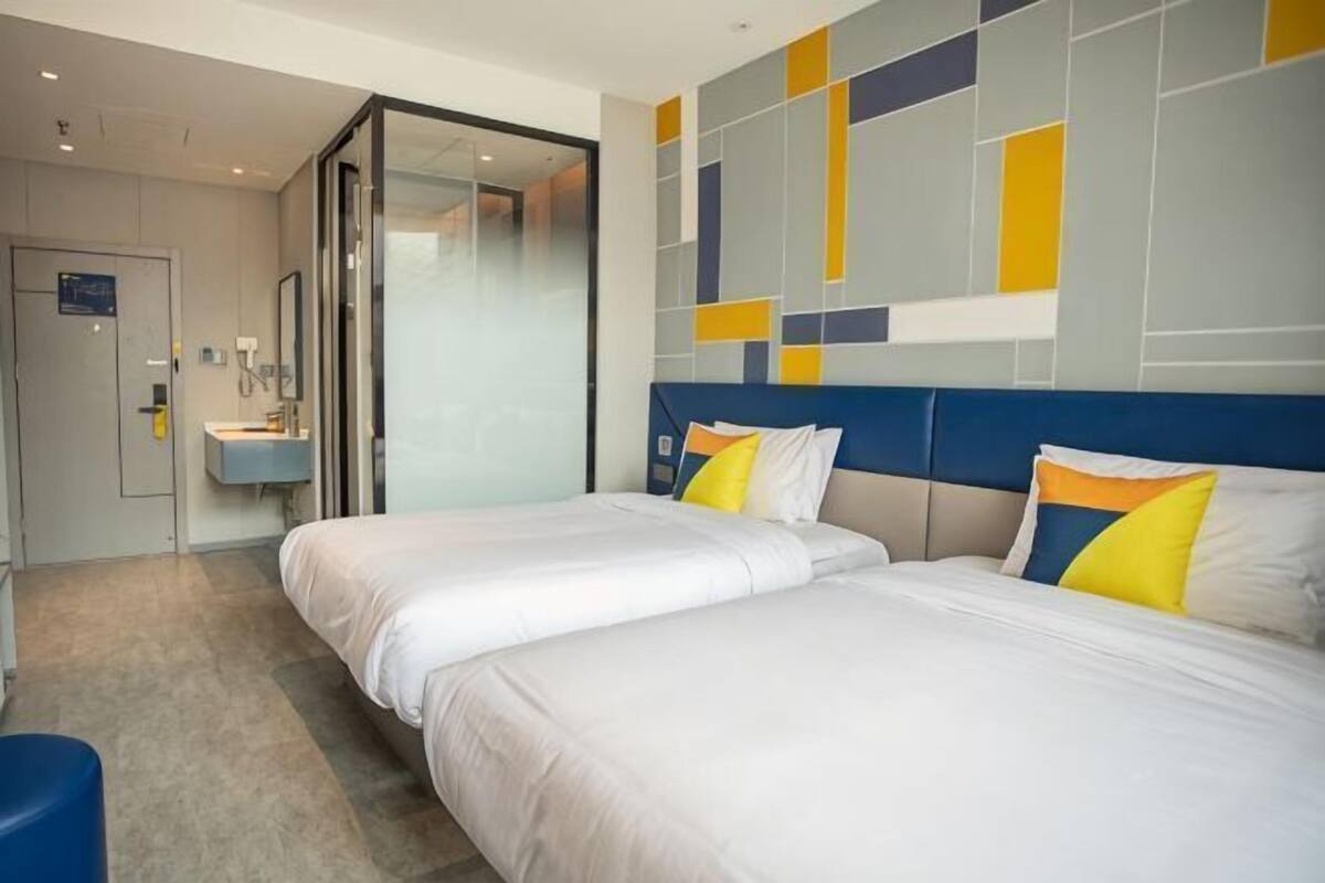 Quarto de hotel com duas camas, banheiro com pia e porta de vidro que dá para outro cômodo.