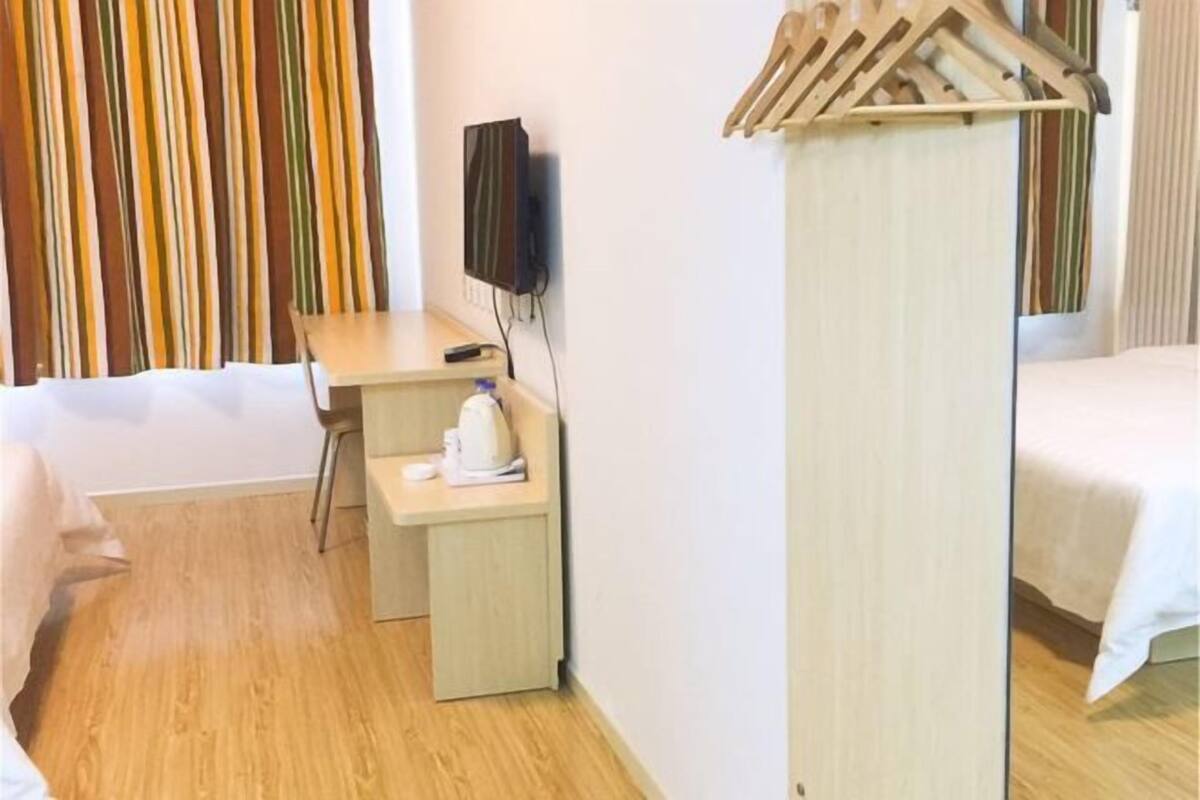 Quarto de hotel com cama, escrivaninha com cadeira, televisão e cabideiro de madeira.