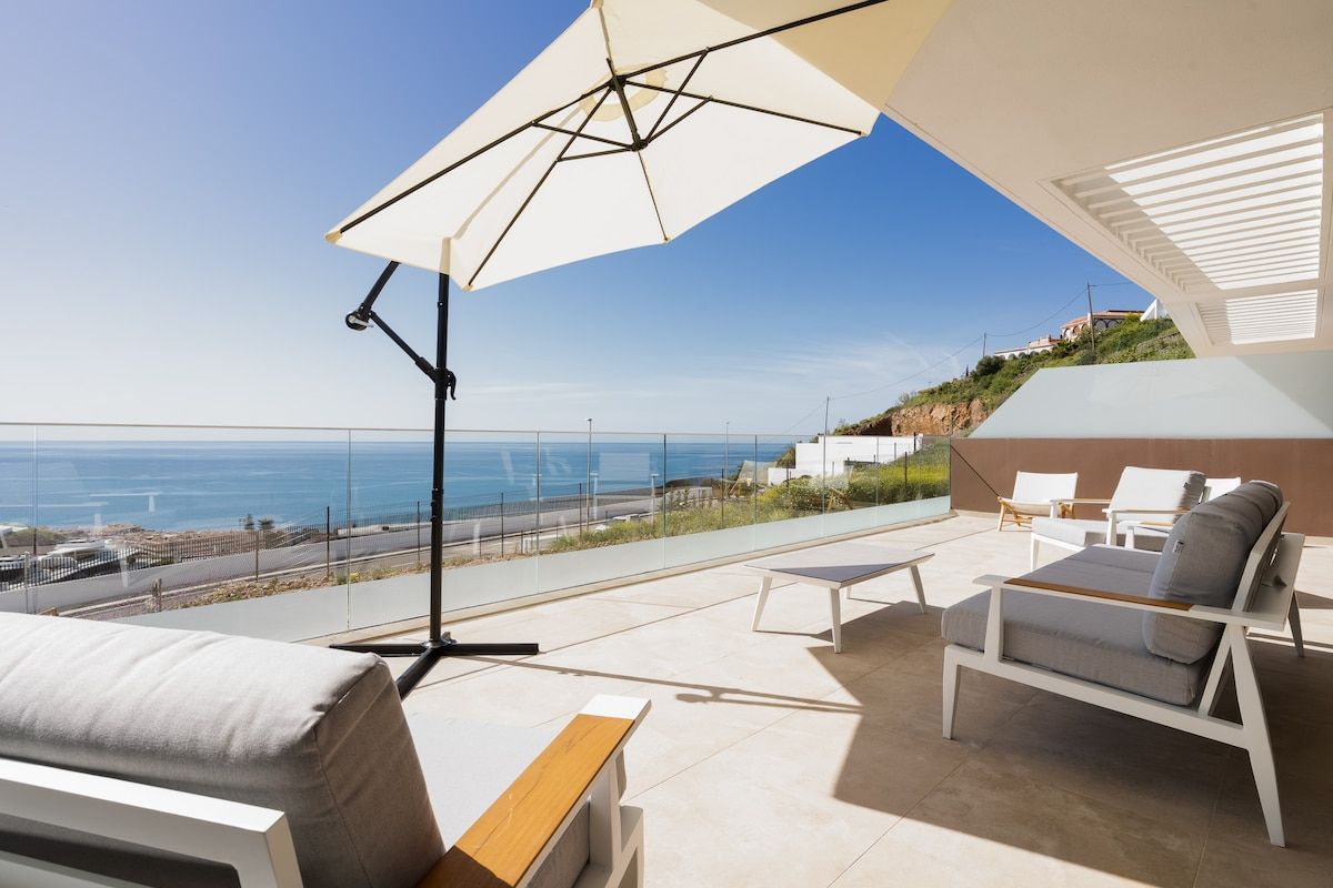 Een terras met uitzicht op zee, buitenmeubels en een grote parasol.
