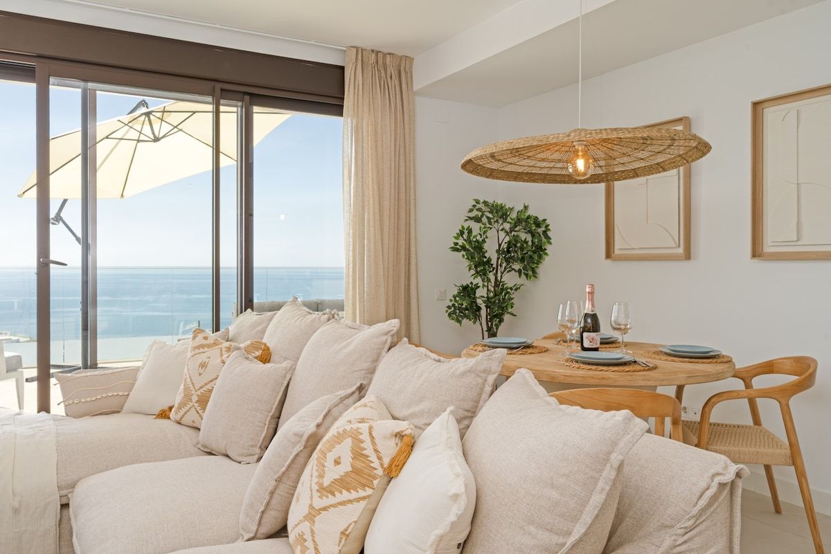 A modern living room with a large window, a round dining table with chairs, and a hanging light fixture.