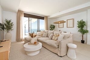 A modern living room with a large sofa, a coffee table, and a view of the sea through a sliding glass door.
