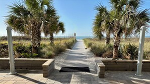 Ein gepflasterter Weg führt zu einem Strand, gesäumt von Palmen und Pflanzen, mit freiem Blick auf den Ozean.