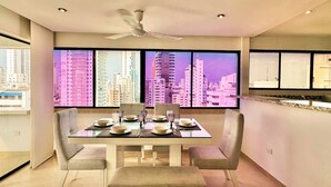 Un espace repas moderne avec une table en verre, quatre chaises et une vue sur le paysage urbain grâce à de grandes fenêtres.