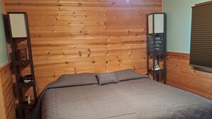 Une chambre avec un plafond en bois, un lit recouvert d’une couette grise, un ventilateur de plafond et deux tables de chevet avec des lampes et des livres.