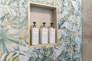 A bathroom with a tiled wall featuring a tropical leaf pattern. There is a shelf with three bottles labeled shampoo, body wash, and conditioner. - Stylish 1BR/1.5Bath Near Liberty Bell (Philadelphia)