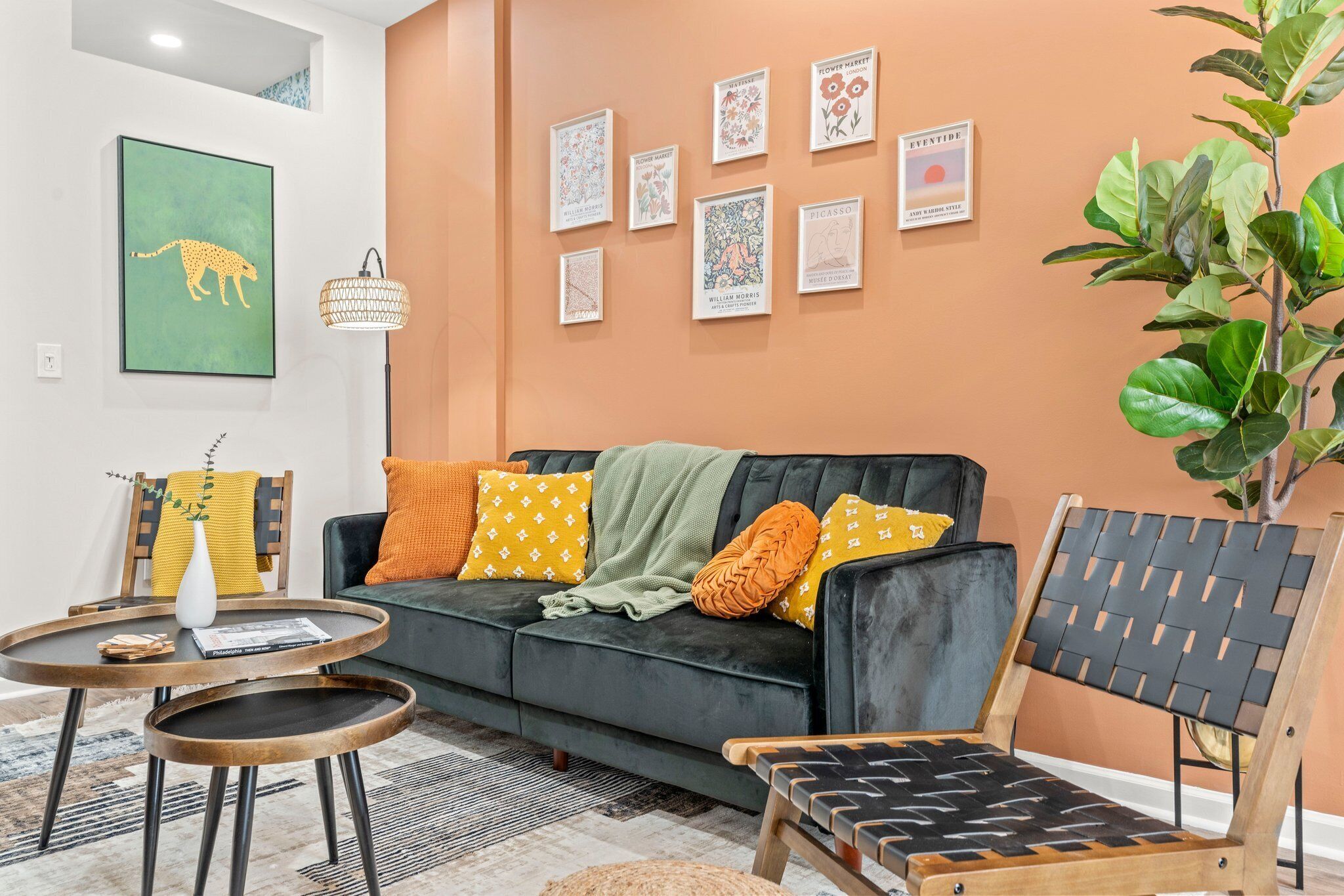 A living room with a black sofa, a round coffee table, and a wooden chair. There are framed artworks on the wall and a large plant in the corner.