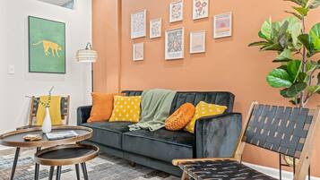A living room with a black sofa, a round coffee table, and a wooden chair. There are framed artworks on the wall and a large plant in the corner.
