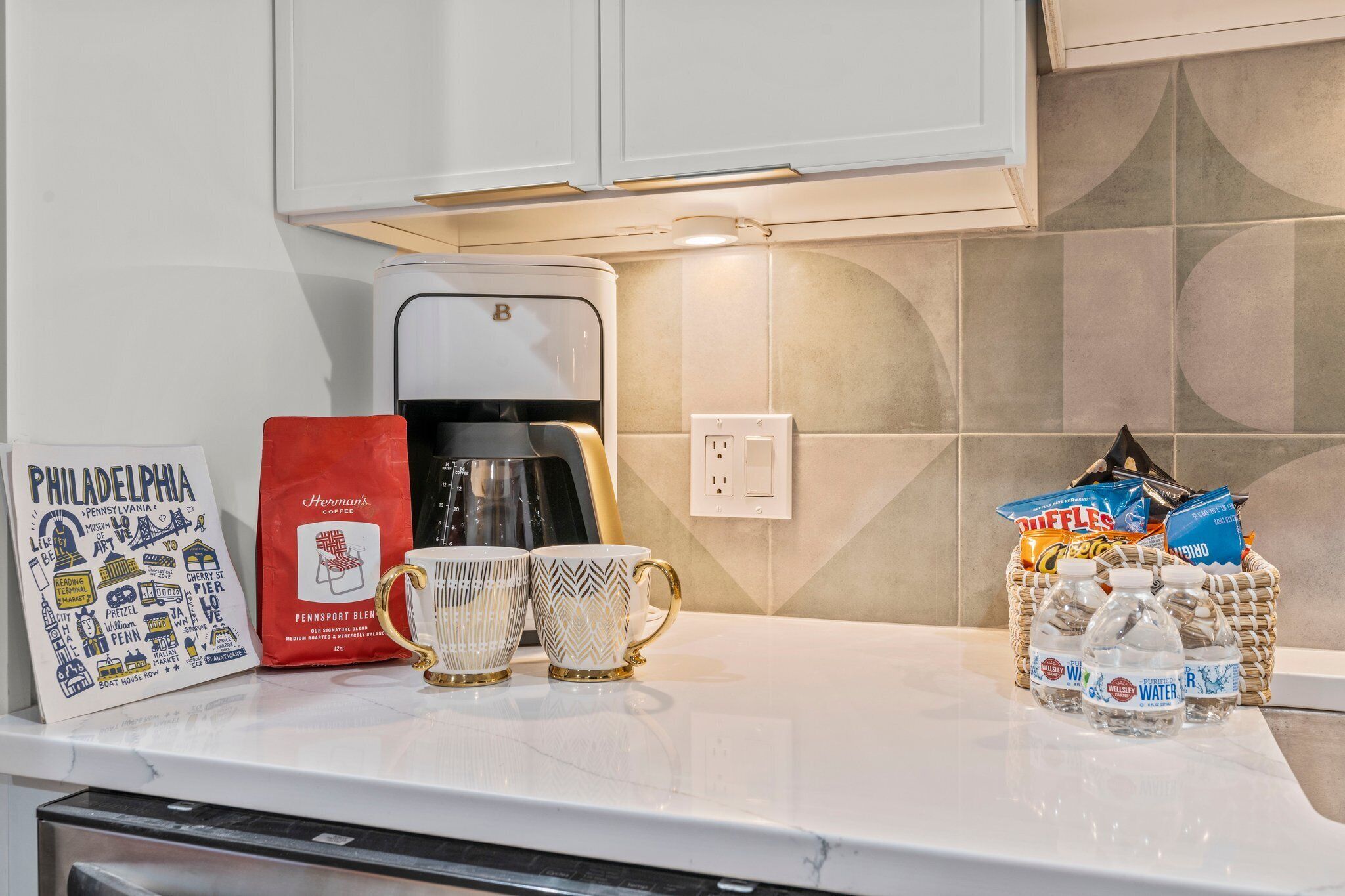 A kitchen counter with a coffee maker, a coffee bag, two mugs, and a basket of bottled water.