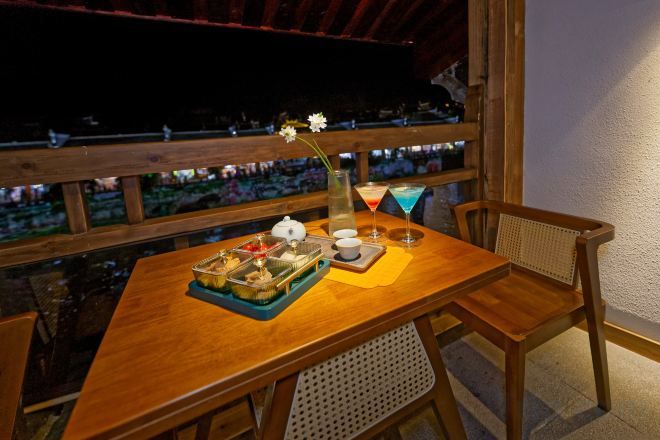 A wooden dining table with a tray of food, a vase with a flower, and a drink on a balcony with a view.