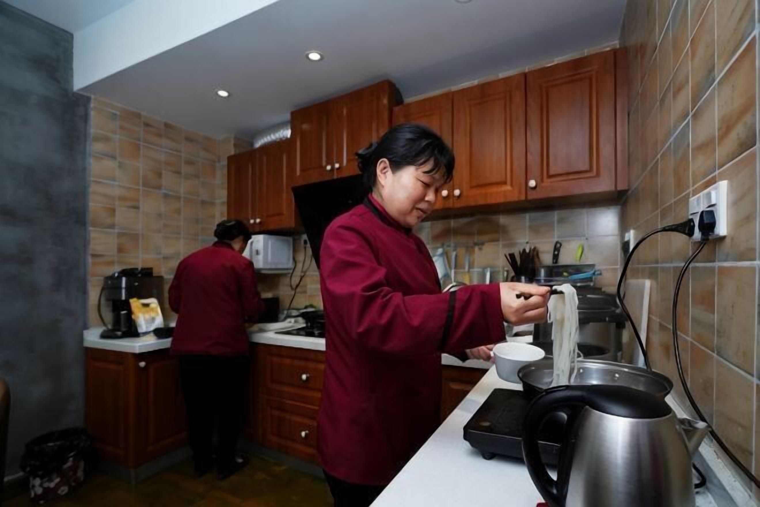 A kitchen with wooden cabinets, a countertop, a kettle, and a person preparing food.