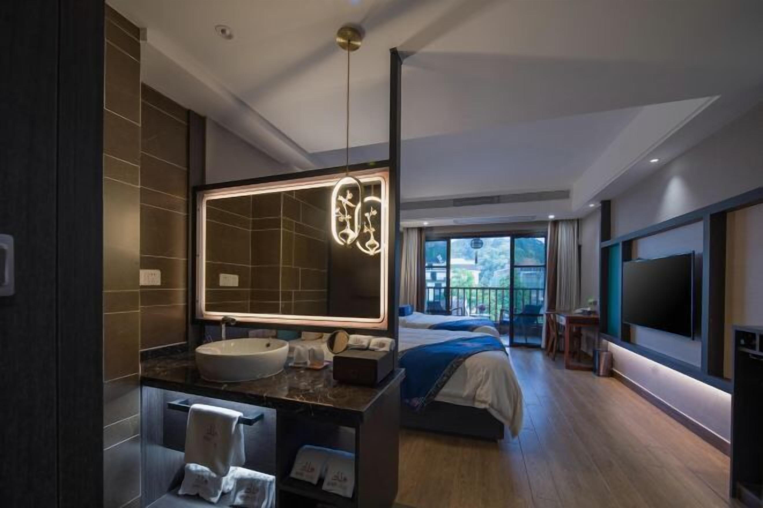 A modern hotel room with a large mirror, a round sink, a bed with a blue blanket, a TV mounted on the wall, and a balcony with a view.
